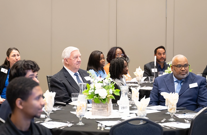 A group of formally dressed people sit at a table, listening to a speech.