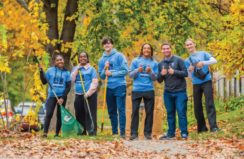 Six college students in blue “Bee the Difference SAU 2024” shirts smile and pose with rakes among fall leaves and colorful trees.
