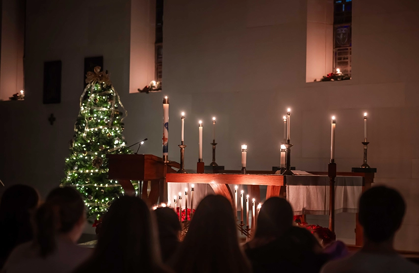 A crowd sits quietly as a candlelight and a christmas tree glitter.