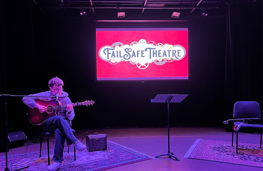 A student plays guitar on stage with a pink screen that reads 