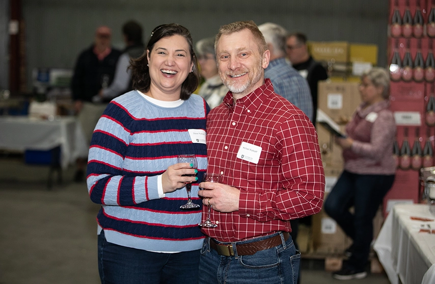 Two participants at the SAU wine at the warehouse stand together and smile at the camera while holding glasses of wine.