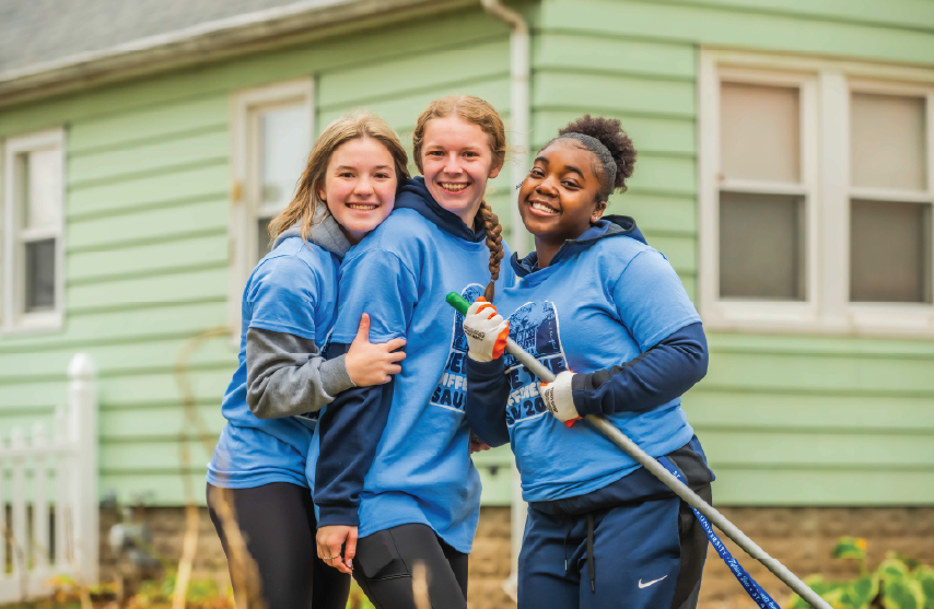 three female students wearing matching blue shirts smile together outdoors while volunteering, holding yard tools in front of a house.