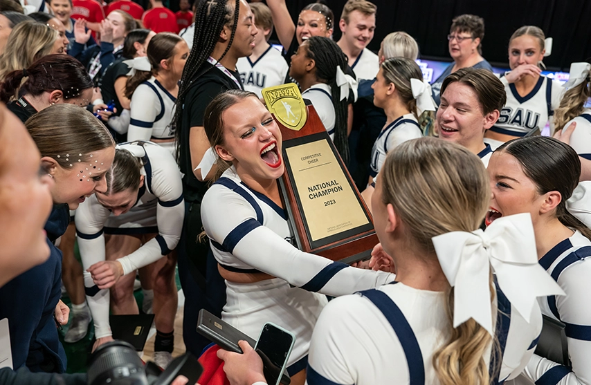 St. Ambrose University's Cheer team smiles in excitement while holding their 2023 First Place trophy at NAIA tournament.