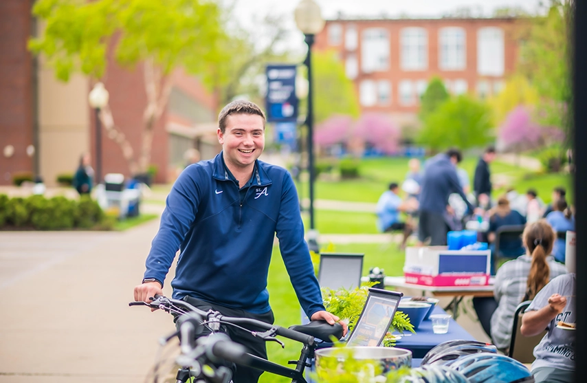 A student smiles at the camera during a club fair where he leans against a bike.