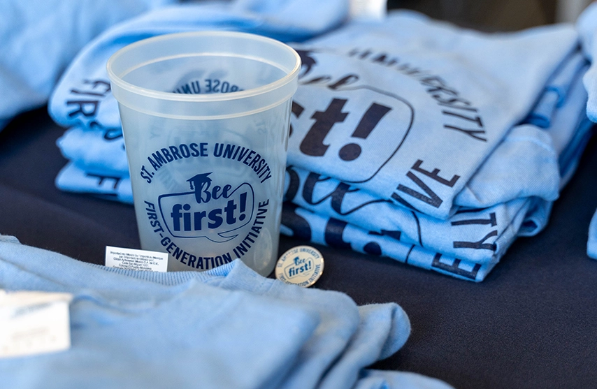 St. Ambrose Branded plastic cup sits among St. Ambrose branded blue shirts.