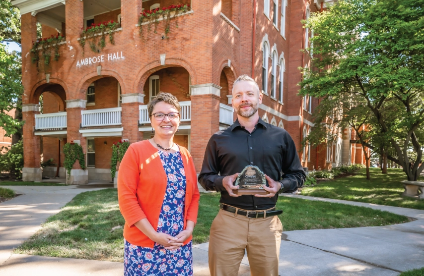 Two people posing for a picture outside of a brick building holding an award.