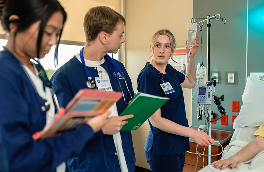 Students of St. Ambrose University's nursing program gather around a hospital bed with folders and notebooks.