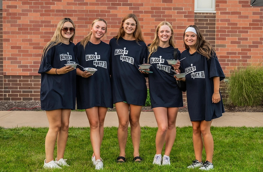 A group of students in matching Block Party Ambrose shirts pose together for a photo outside while holding bowls of ice cream.