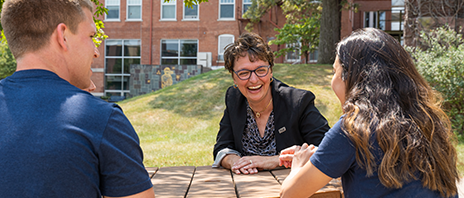 Amy Novak, EdD sits a table and laughs with two students dressed in dark blue.