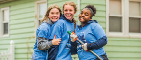 three female students wearing matching blue shirts smile together outdoors while volunteering, holding yard tools in front of a house.