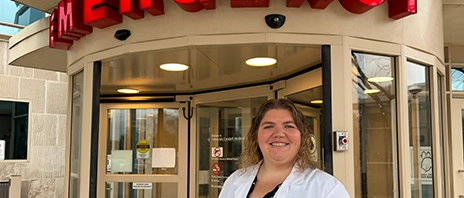 Ashley Vilona, '14, smiles in her doctor's coat while standing in front of Emergency Room revolving doors.
