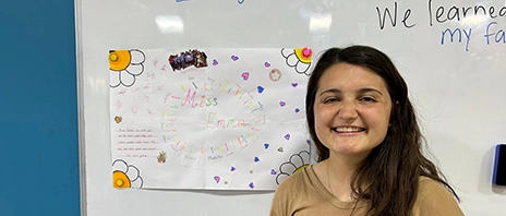 Emma Janecek stands in front of a colorful white board and smiles at the camera.