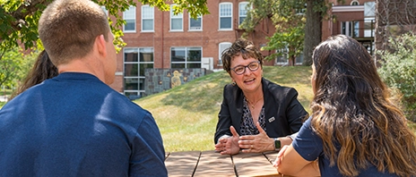 President Amy Novak, EdD, of St. Ambrose University sits and talks to students at a round wooden table outside on St. Ambrose University's campus.