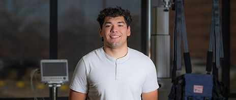 Oscar Mejia, '27 DPT, stands in an physical therapy lab at St. Ambrose University and smiles into the camera.
