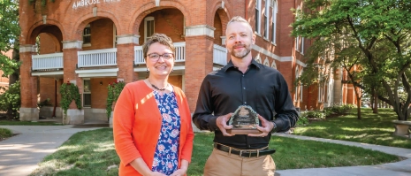 Two people posing for a picture outside of a brick building holding an award.