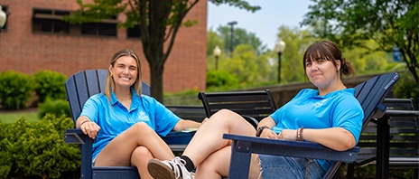 Students smile at the camera as they relax in two SAU blue adirondack chairs.