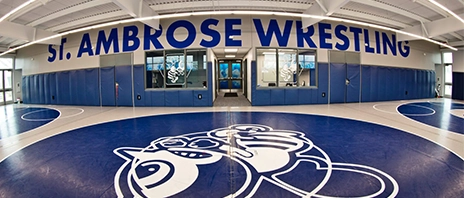 The blue and white wrestling gym at SAU with the SAU Bee on the floor and blue St. Ambrose Wrestling on  the rafters.