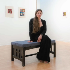 An art student sitting on a bench in a gallery. The exibit around her features her artwork.