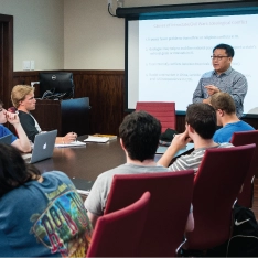 A group of students sitting around a boardroom style room during a presentation.