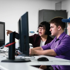 Students focused on computers in a classroom with their professor pointing something out on the screen.