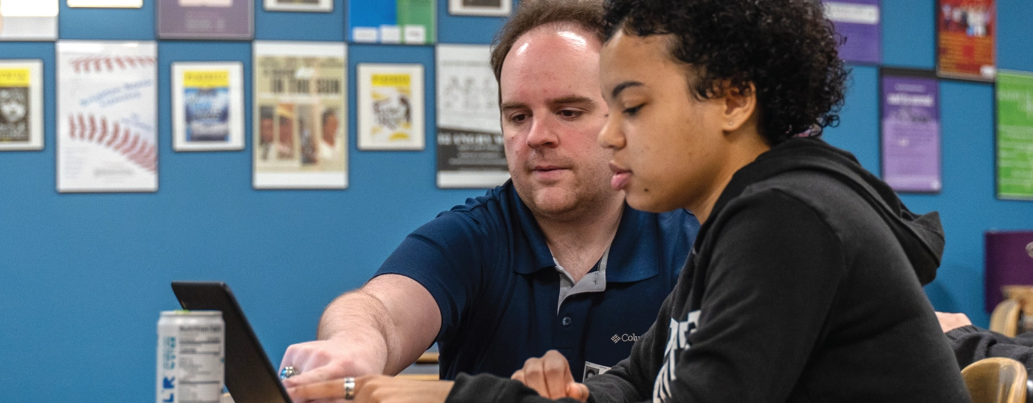 A student teacher pointing at a computer over the shoulder of a young student.