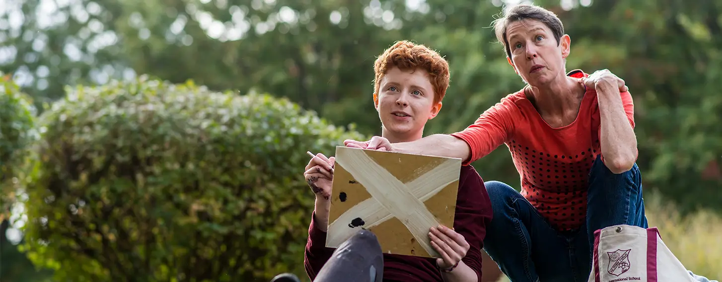 A woman sitting on the grass with a student who is holding a painting.