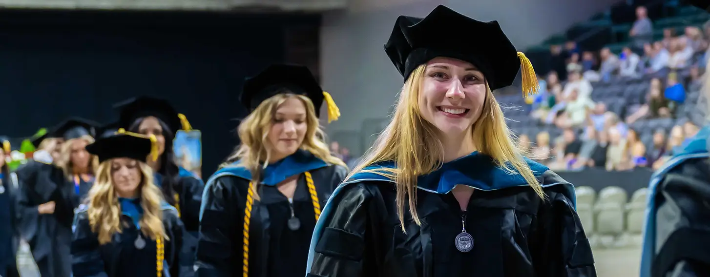 Group of graduates in caps and gowns walking down aisle at commencement ceremony.