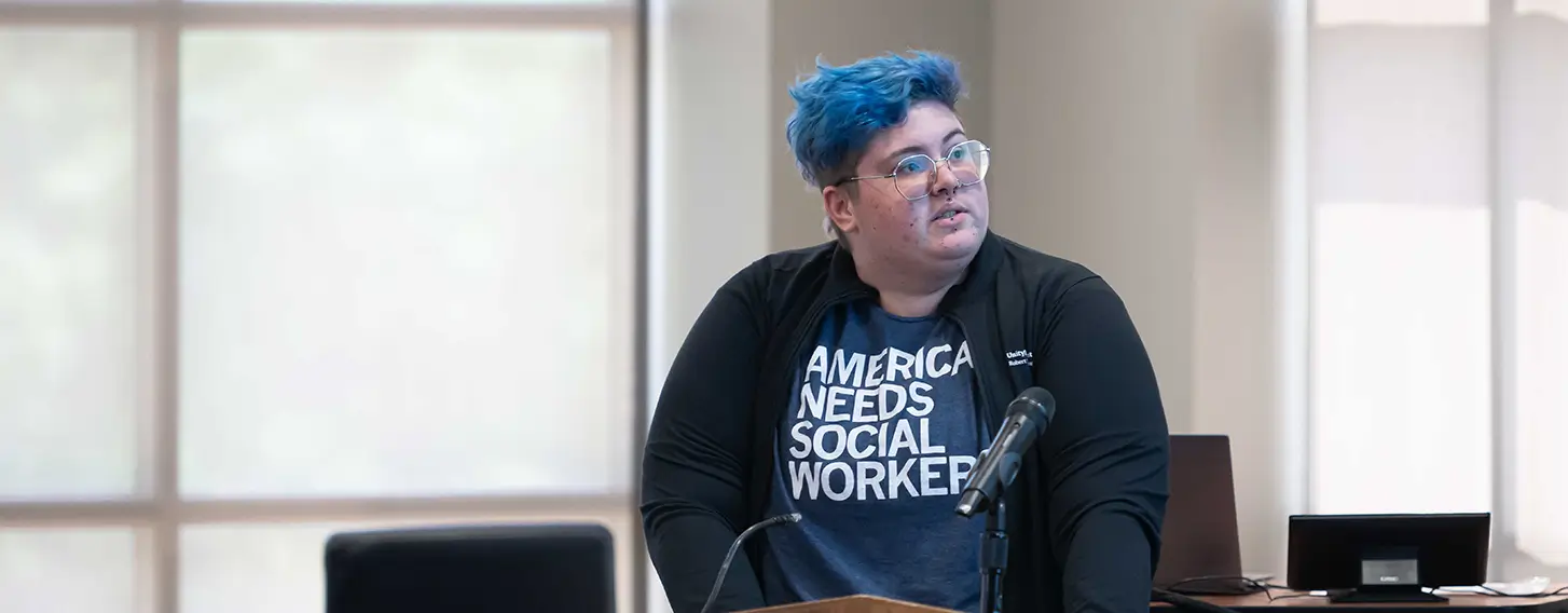 A person with blue hair standing at a podium, addressing an audience.