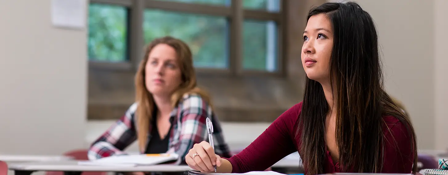 Female students sitting at desks in a classroom with books, taking notes.