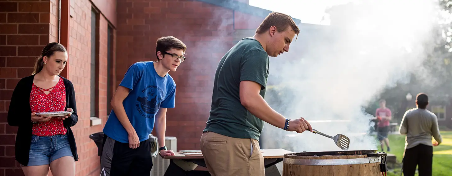 A gathering of students surrounding a barrel smoking food at upperclassmen housing.