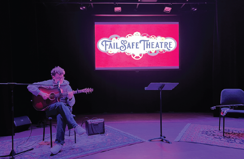 A student sitting on stage playing guitar with a sign that says Fail Safe Theatre