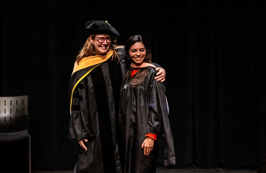 A newly hooded Masters student smiles and stands next to her professor at the St. Ambrose University Master's Hooding Ceremony.