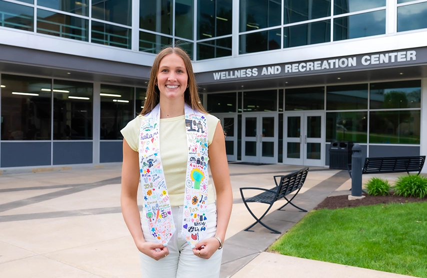 Abby Wolter, '26, smiles into the camera in front of St. Ambrose University's Wellness and Recreation center.