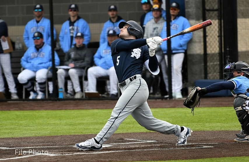 St. Ambrose baseball player follows through on a swing at home plate as teammates watch from the dugout in the background.