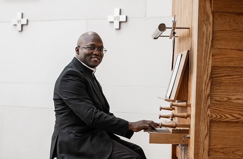 Fr. Eric Koami '25 smiles into the camera as he sits at the piano in Christ the King Chapel at St. Ambrose University.