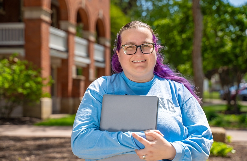 Jennifer Parsons smiles into the camera while holding her laptop on St. Ambrose University's campus.