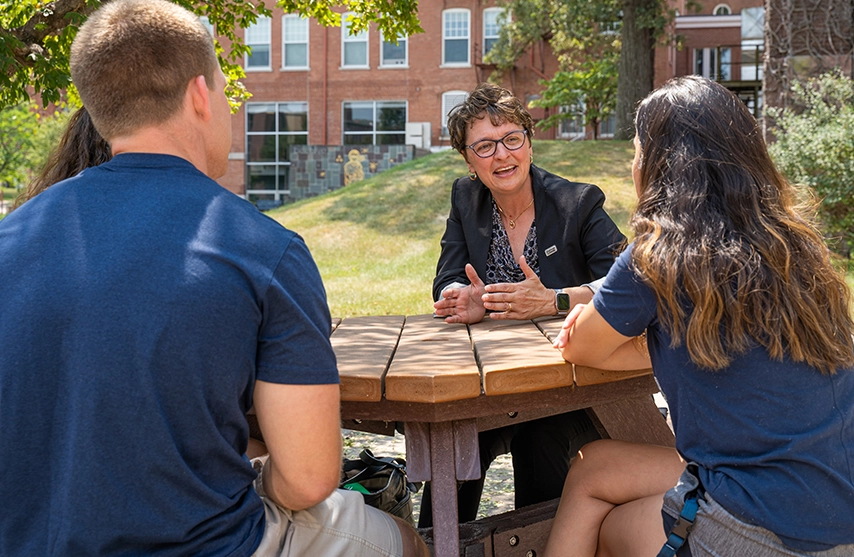 President Amy Novak, EdD, of St. Ambrose University sits and talks to students at a round wooden table outside on St. Ambrose University's campus.