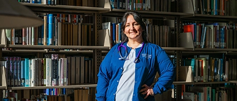 Kathy Franco stands in the St. Ambrose University Library in front of a shelf of books and smiles into the camera as a beam of light illuminates her.