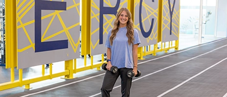 A student stands on an indoor track holding a dumbbell in each hand.